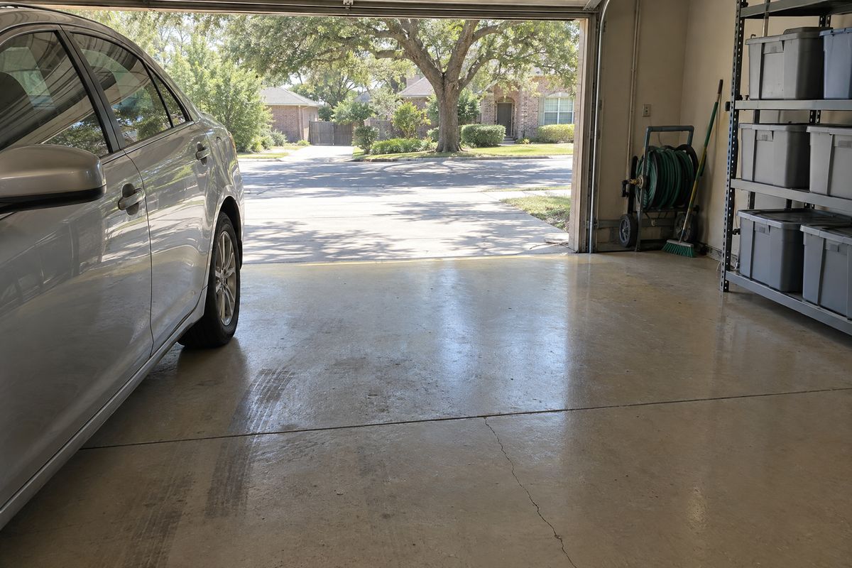 A garage floor in solid tan epoxy with dark tire marks, a hairline crack running from a joint and a sunlit band near the open door, beside a parked sedan