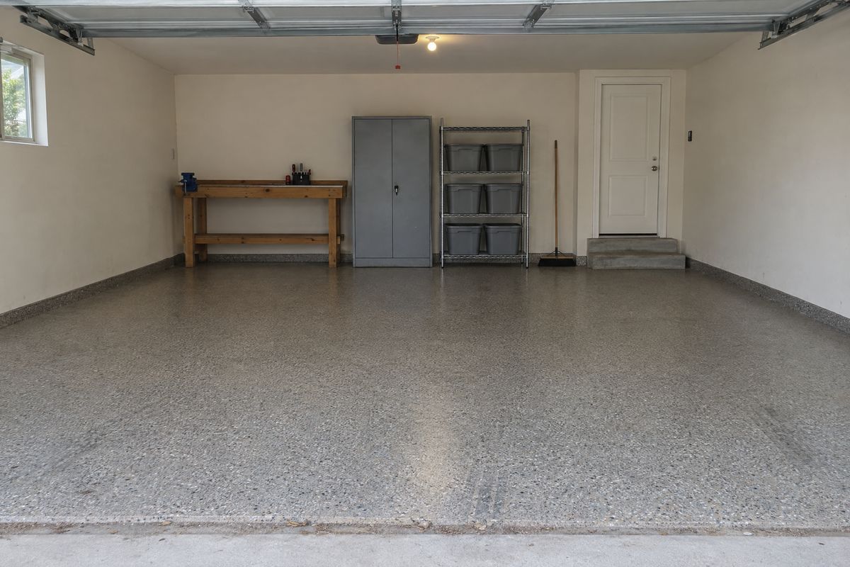 A two-car garage floor covered in gray, tan and black vinyl flakes, with a workbench, wire shelving with gray bins and steps up to a house door