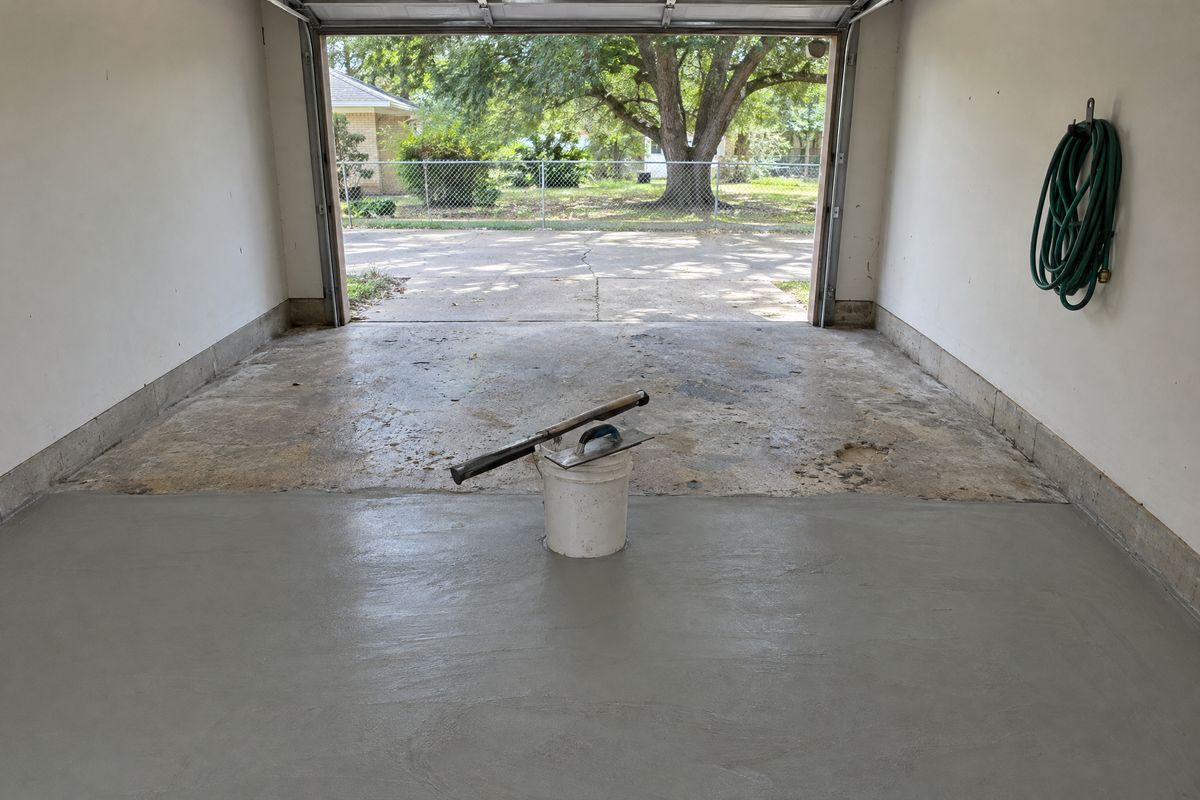 A one-car garage floor where the far half is worn, pitted and oil stained and the near half has a smooth pale gray cement resurfacer with trowel marks