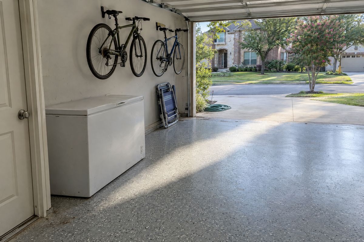 Late afternoon sun crossing a light gray flake garage floor with a satin clear topcoat, with a chest freezer, bicycles on wall hooks and the driveway outside