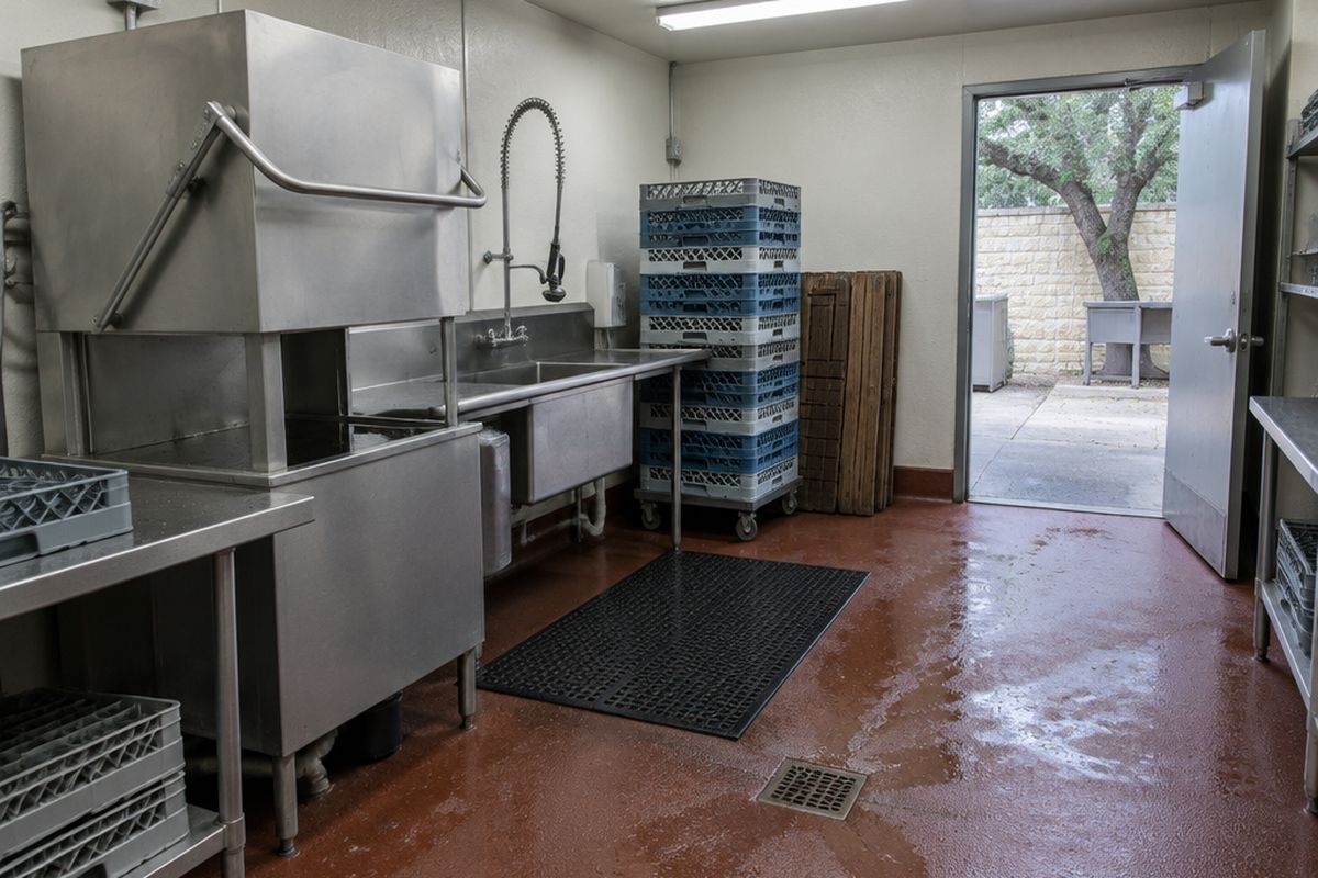 A restaurant dish room with a wet red-brown urethane cement floor sloping to a drain, a stainless dish machine, stacked dish racks and wooden duckboards