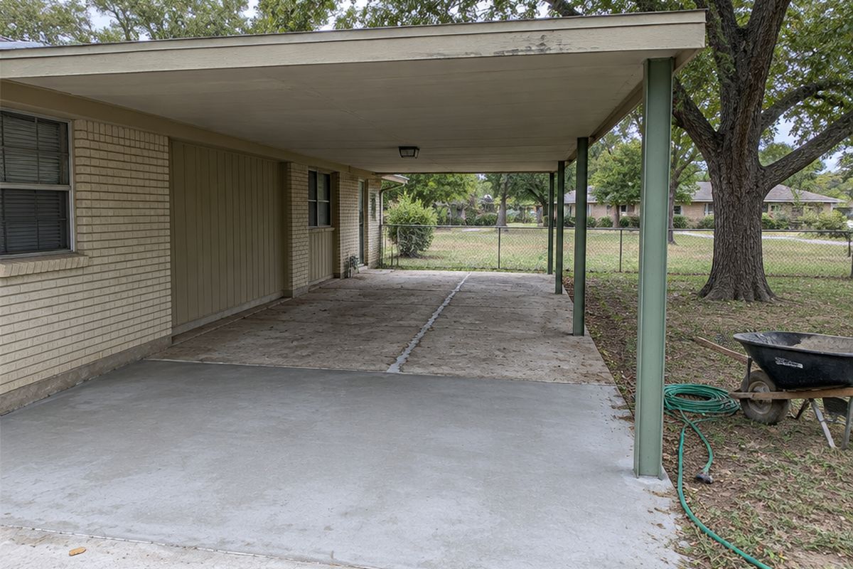 The carport of an older one-story ranch house, its slab half covered in smooth gray resurfacer and half still scaled and pitted, with a wheelbarrow and a pecan tree