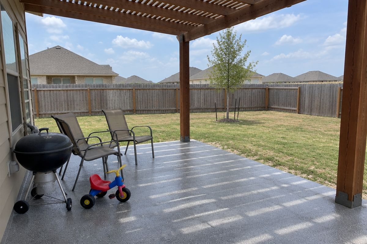 A back patio under a wooden pergola with a gray flake coating, a charcoal grill, two chairs and a child's tricycle, in a flat yard with a privacy fence