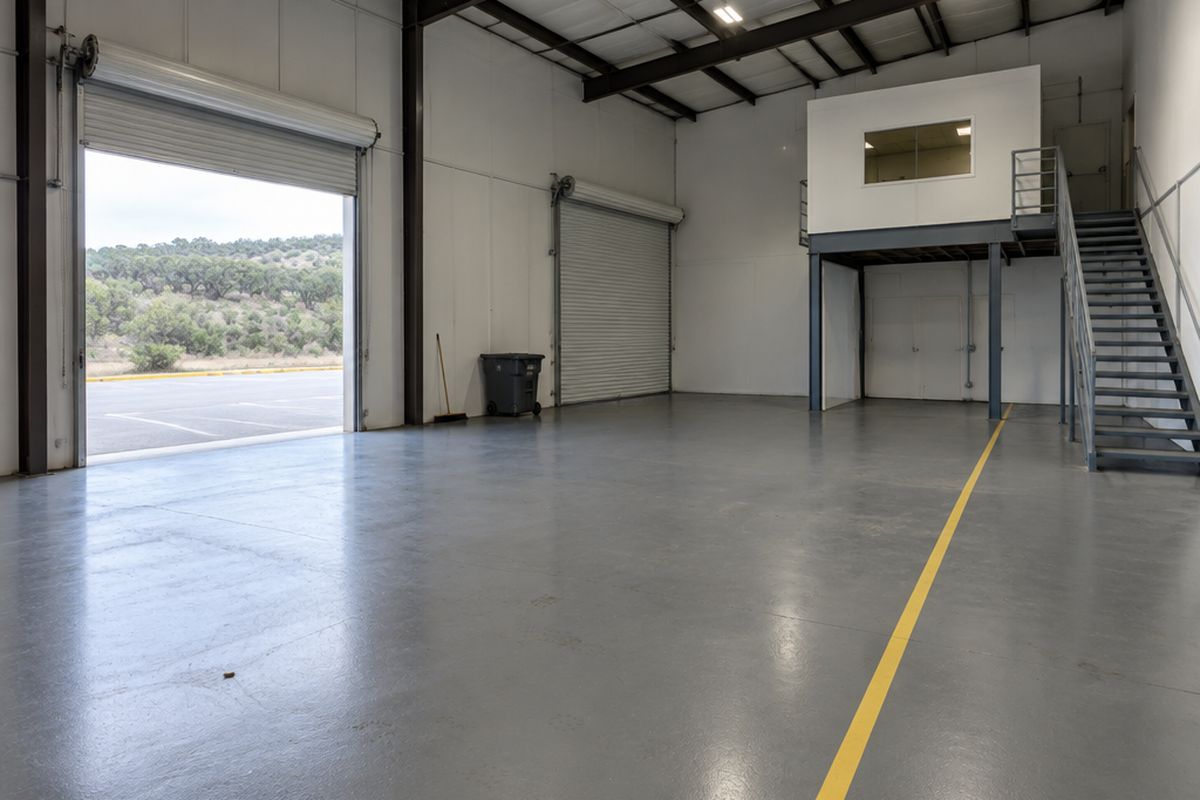 An empty flex warehouse suite with a gray epoxy floor, a yellow walkway line, a mezzanine office with a railing and an open roll-up door to juniper hills