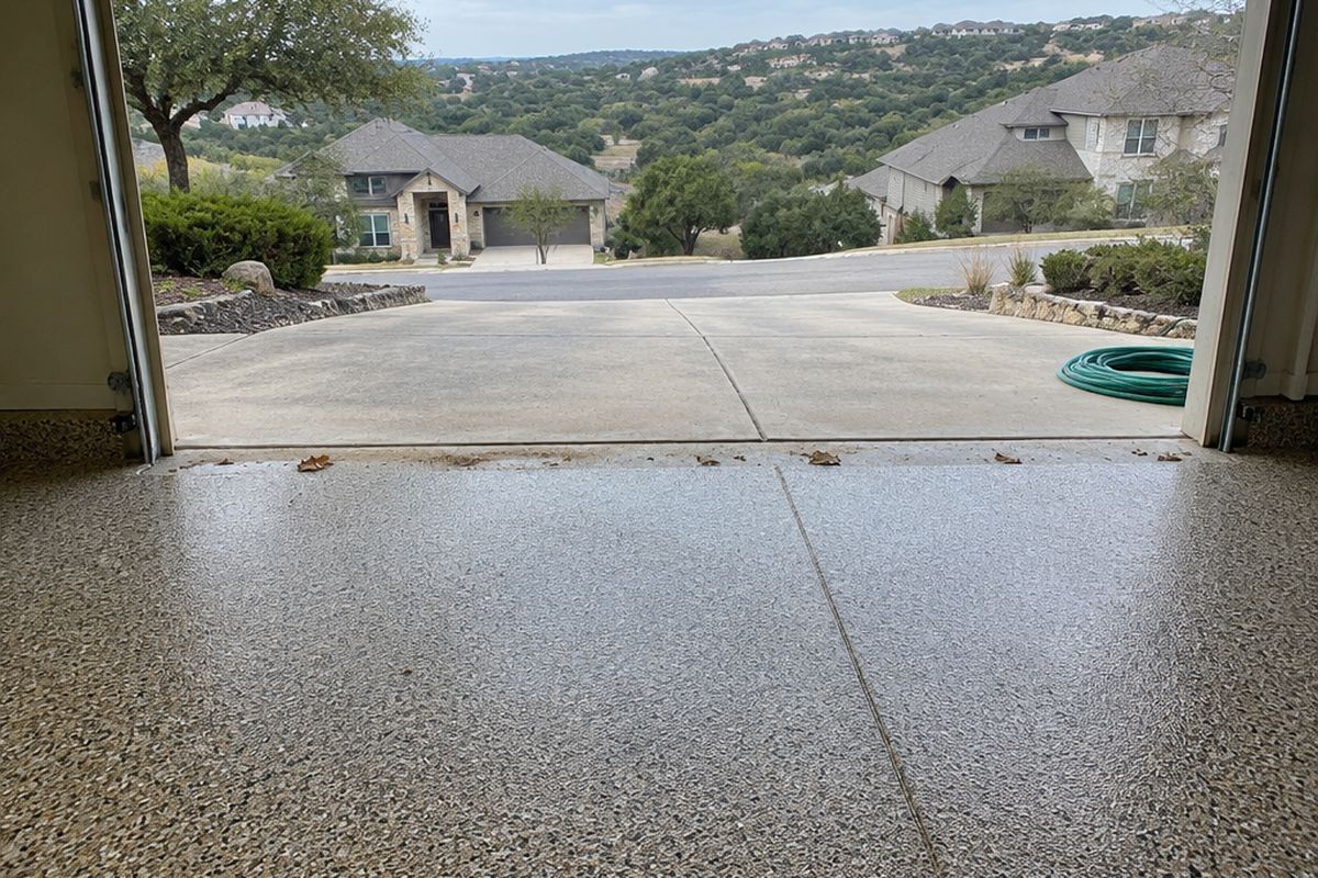 A garage threshold with a tan, gray and black flake floor, a joint line, dust and leaves at the edge, a garden hose and a driveway sloping toward wooded hills
