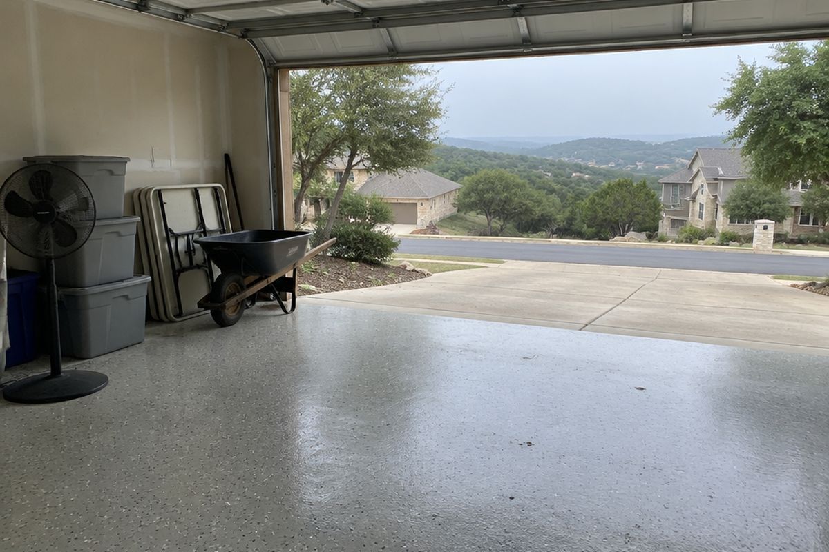 A two-car garage on a humid morning with a light gray flake floor under a satin topcoat, a pedestal fan, plastic bins and a wheelbarrow