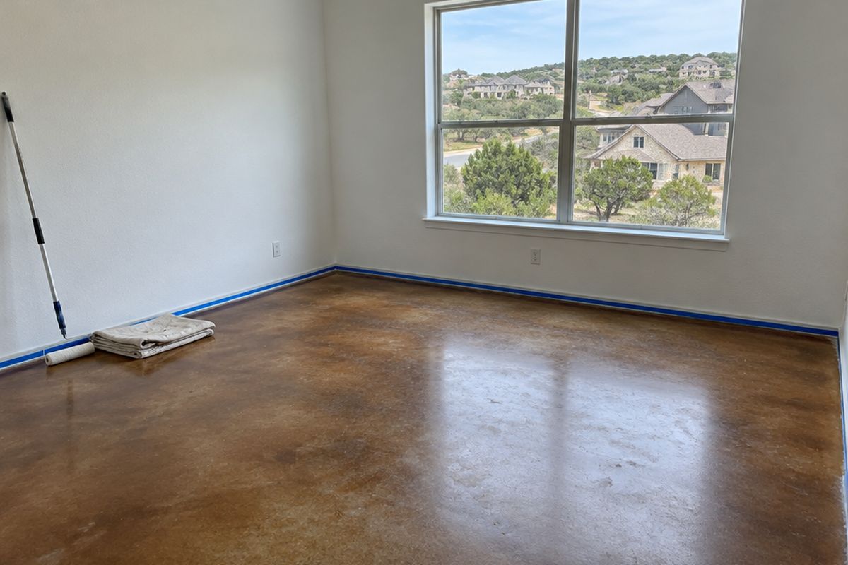 An empty room in a new house with a cola-brown and tan stained concrete floor, painter's tape on the baseboards, a paint roller and a folded drop cloth