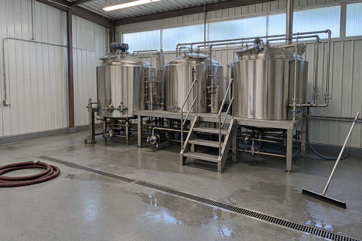 A brewhouse with three stainless brewing vessels on a low platform, a gray urethane cement floor, a stainless trench drain, a coiled hose and puddles